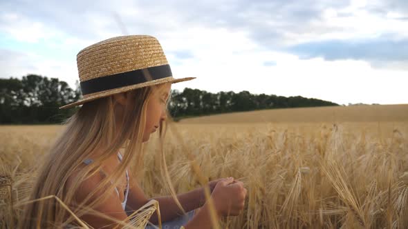 Happy Small Child in Straw Hat Holding Cereal Spikelet in Hands While Sitting in the Wheat Field