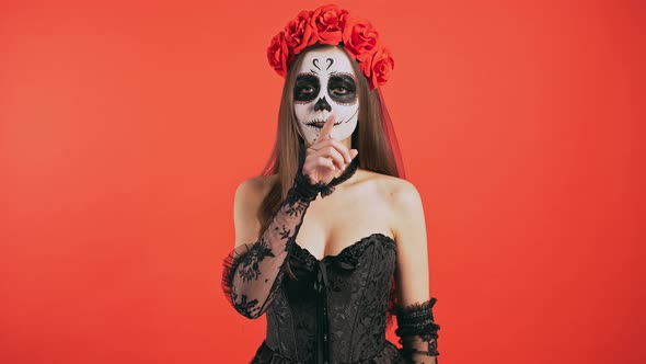 Day of Dead Woman with Sugar Skull Makeup is Smiling Showing Be Quiet Sign Posing on Red Background