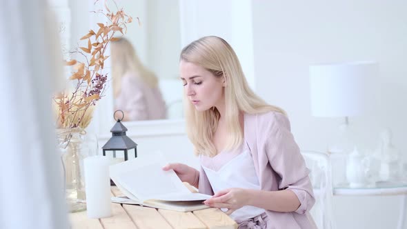 Young Beautiful Woman Reads a Book at the Table in the Living Room