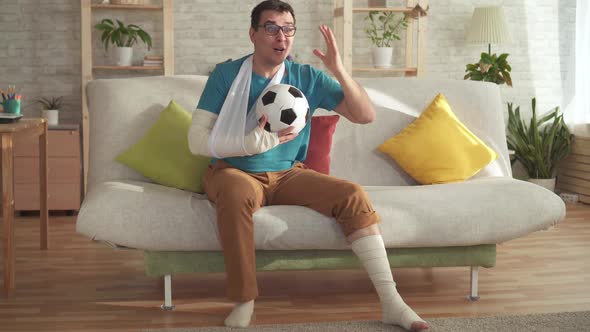 Expressive Young Man with a Broken Arm and Leg Sitting on the Couch with a Soccer Ball Learns About