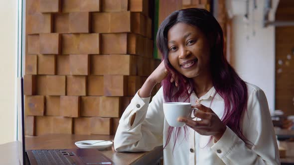 Young African Woman Drinking Coffee and Working on a Laptop While Sitting in a Cafe