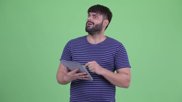 Happy Young Overweight Bearded Indian Man Thinking While Using Digital Tablet