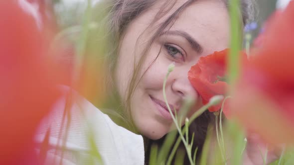 Portrait of Cute Young Woman Sitting in Poppy Field Looking at Camera