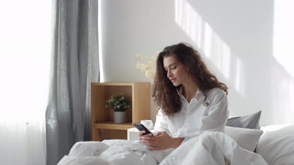 Pretty Woman in Headphones Watching Video on Smartphone in Bed