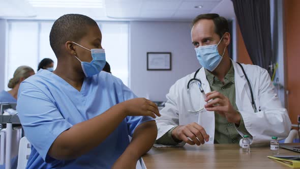 Caucasian male doctor giving covid vaccination to african american female doctor, both in face masks