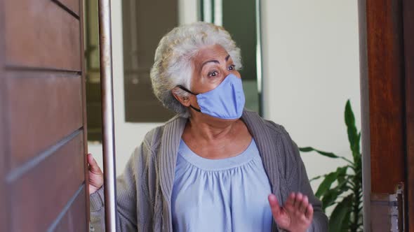 African american senior woman wearing face mask opening front door and welcoming at home