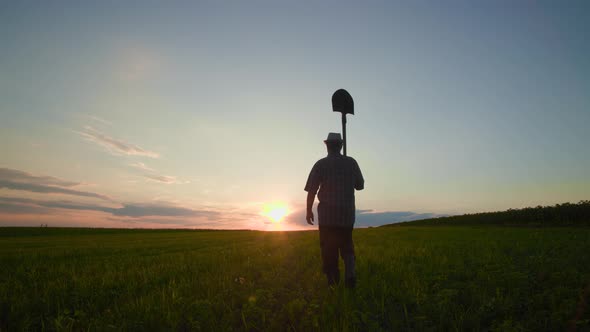 Silhouette of a Male Farmer Working in the Field at Sunset with a Shovel Walks Through Farmland
