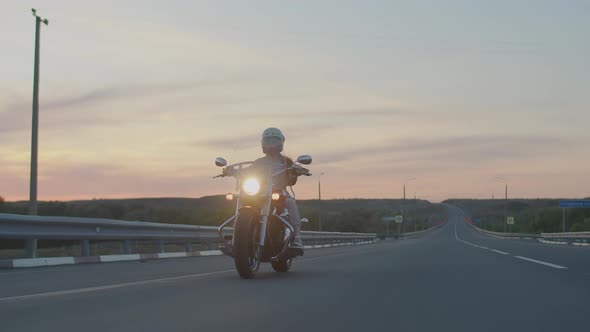 Woman in White Helmet on Motorcycle with Headlights on is Driving Along Evening Highway Front View