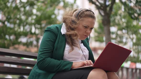 Portrait of Confused Plussize Woman in Earphones Sitting on Bench Outdoors with Laptop Waving at