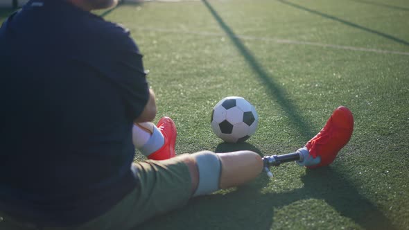 Zoom in to Soccer Ball on Green Grass with Unrecognizable Male Amputee Sitting on Stadium