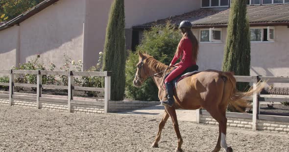 Caucasian Girl in Pink Clothes and Helmet Riding Brown Horse in the Corral. Young Female Equestrian