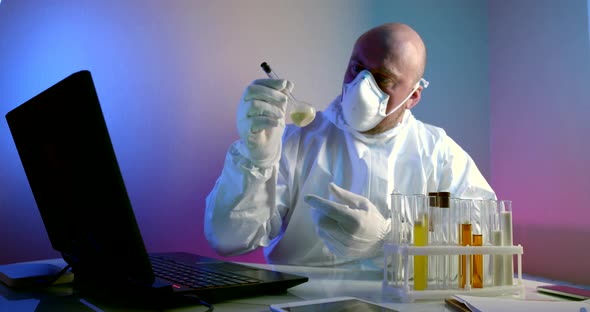 Virologist in Protective Clothing Studies the Substance in a Test Tube Sitting at a Table