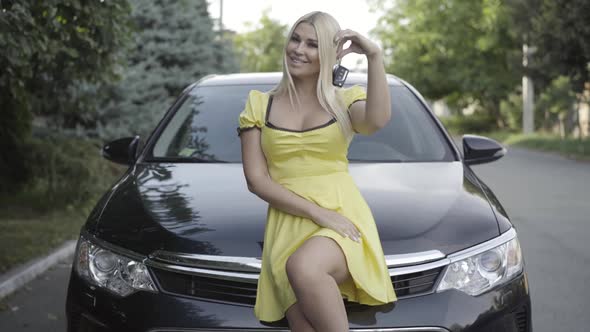 Portrait of Attractive Young Woman Sitting on Car Hood and Showing Keys. Happy New Owner Posing with