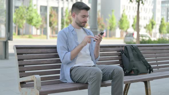 Young Man Browsing Internet on Smartphone While Sitting on Bench