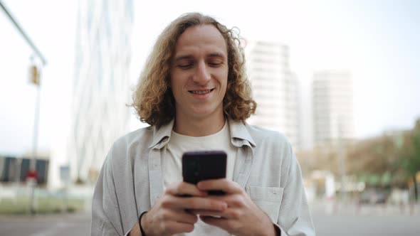 Cheerful curly-haired man typing by mobile
