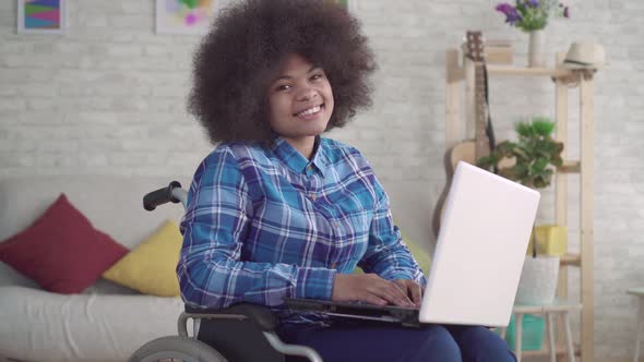 Disabled African American Woman with an Afro Hairstyle in a Wheelchair Uses a Laptop Looking at the