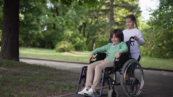 Wide Shot Smiling Disabled Boy with Girl Pushing Wheelchair in Slow Motion Outdoors