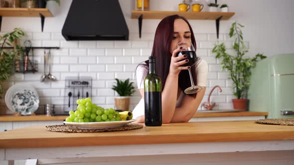Relaxed Young Woman Drinks Red Wine Standing at Kitchen Table