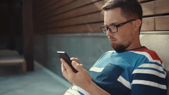 Bearded Man Is Typing on Screen of Smartphone, Lying on Lounger in Sunny Day