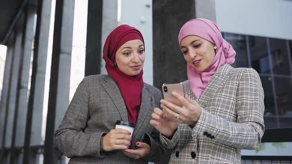 Two Young Muslim Women Wearing Hijab Headscarf Look at Phone, Walking Together Laughing Smile To