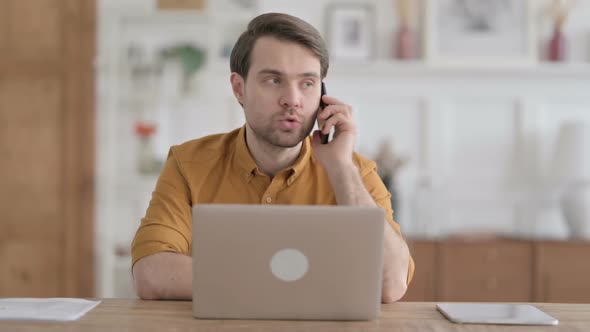 Angry Young Man Talking on Phone while using Laptop in Office