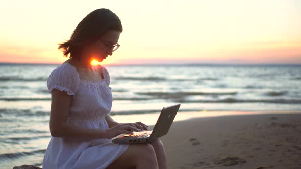 A Young Woman Sits on a Stone on the Beach By the Sea with a Laptop in Her Hands. A Girl in a White