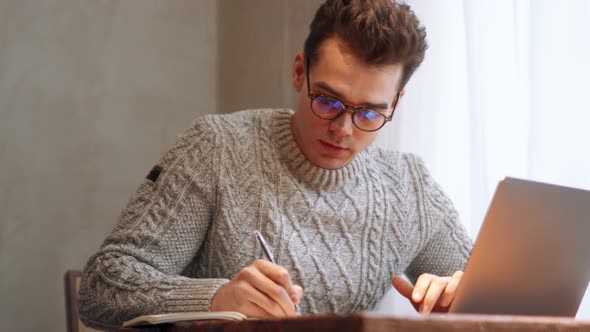 Smiling man wearing eyeglasses writing something from laptop to notepad