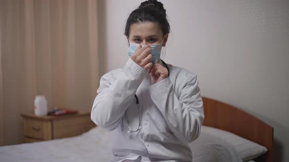 Concentrated Intelligent Young Woman in Medical Gown Putting on Covid Face Mask Waving Hello in