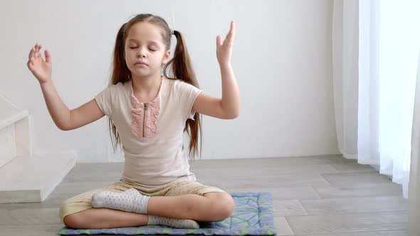 Little Cute Girl Sitting in Yoga Lotus Pose and Meditates Near the Window