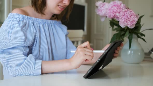 Young Cheerful Woman Working on the Tablet with Pencil in Modern Interior with Peonies on the