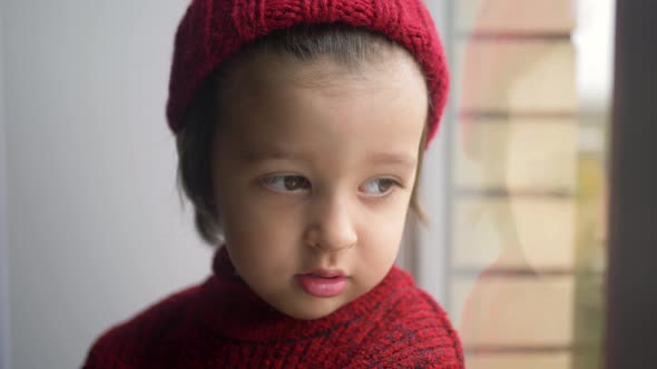 Boy Child in a Red Sweater and a Knitted Hat Sits on a Large White Window