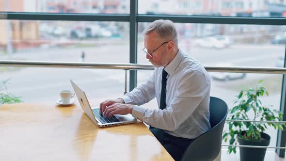 Mature man in white shirt sitting at the table near the window and working on a laptop. 