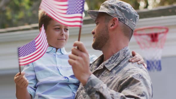 Happy caucasian male soldier carrying his smiling son holding flag in garden outside their house