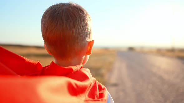 Back View Boy is Standing in Field on Country Road and Behind Him Flutters Red Fabric of Raincoat of