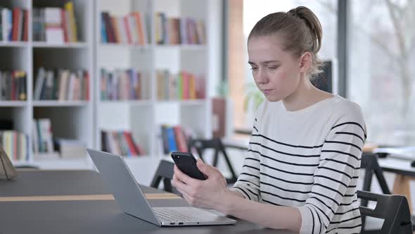 Smartphone Use By Young Woman with Laptop in Library 