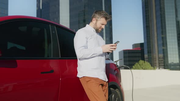 American Businessman Using Smartphone and Leans on Charging Luxury Electric Car