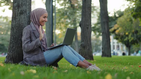 Happy Muslim Islamic Girl Business Woman in Hijab Sitting Near Tree on Green Grass in City Park