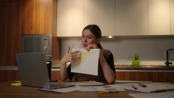 Home Office A Young Woman Shows a Graph in a Laptop Camera Sitting at Home in the Kitchen Conducting