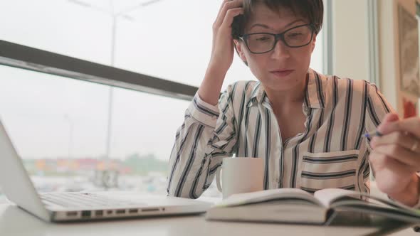 Serious Business Woman in Eyeglasses Works with Laptop and Paper Organiser in Coworking Center