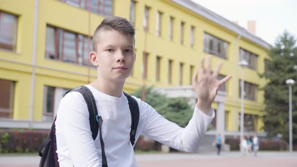 A Caucasian Teenage Boy Waves at the Camera with a Smile  a School in the Background