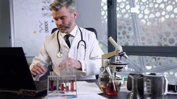 Male Doctor with Gray Beard and Hair Wearing White Gown Working with Microscope and Laptop