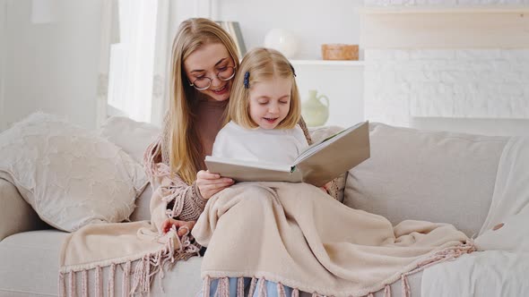 Young Mom Blonde Mother Sits with Little Daughter Baby Girl on Couch Covered By Warm Blanket Reading