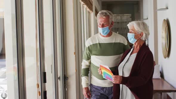 Senior caucasian couple wearing face masks holding rainbow painting against the window at home