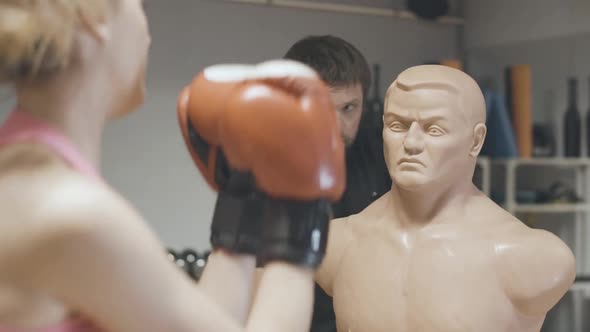 Back View of Blond Female Boxer Punching Mannequin with Blurred Personal Trainer Talking at the