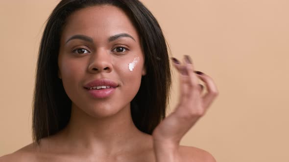 African American Female Applying Moisturizer On Face Over Beige Background