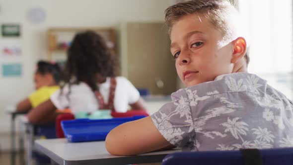 Portrait of happy caucasian schoolboy sitting at classroom, making notes, looking at camera