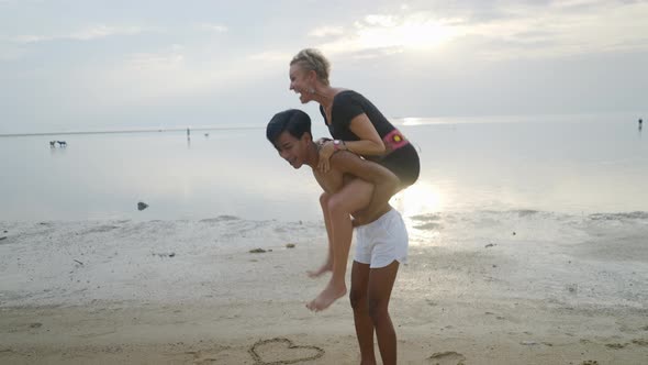 Young Thai Man and Mature Caucasian Woman Couple Piggyback Ride at the Beach at Sunset