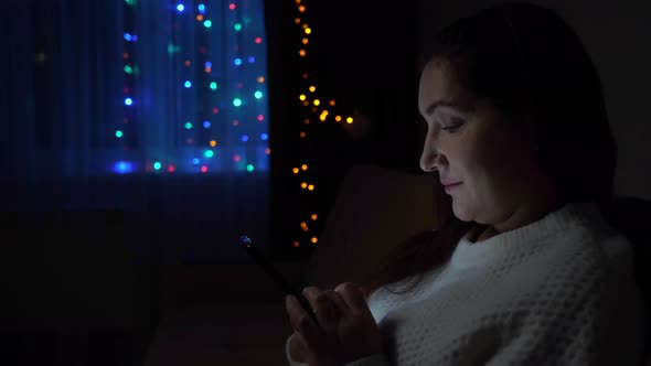 Young Woman Scrolls Social Media Sitting on Sofa at Darkness