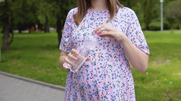 A Pregnant Woman in a Summer Dress with a Floral Print Stands in the Park and Drinks Pure Spring