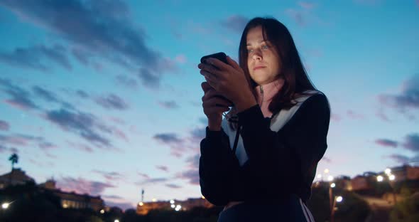 Smiling Young Attractive Woman Uses Smartphone in Park on Evening Sunset